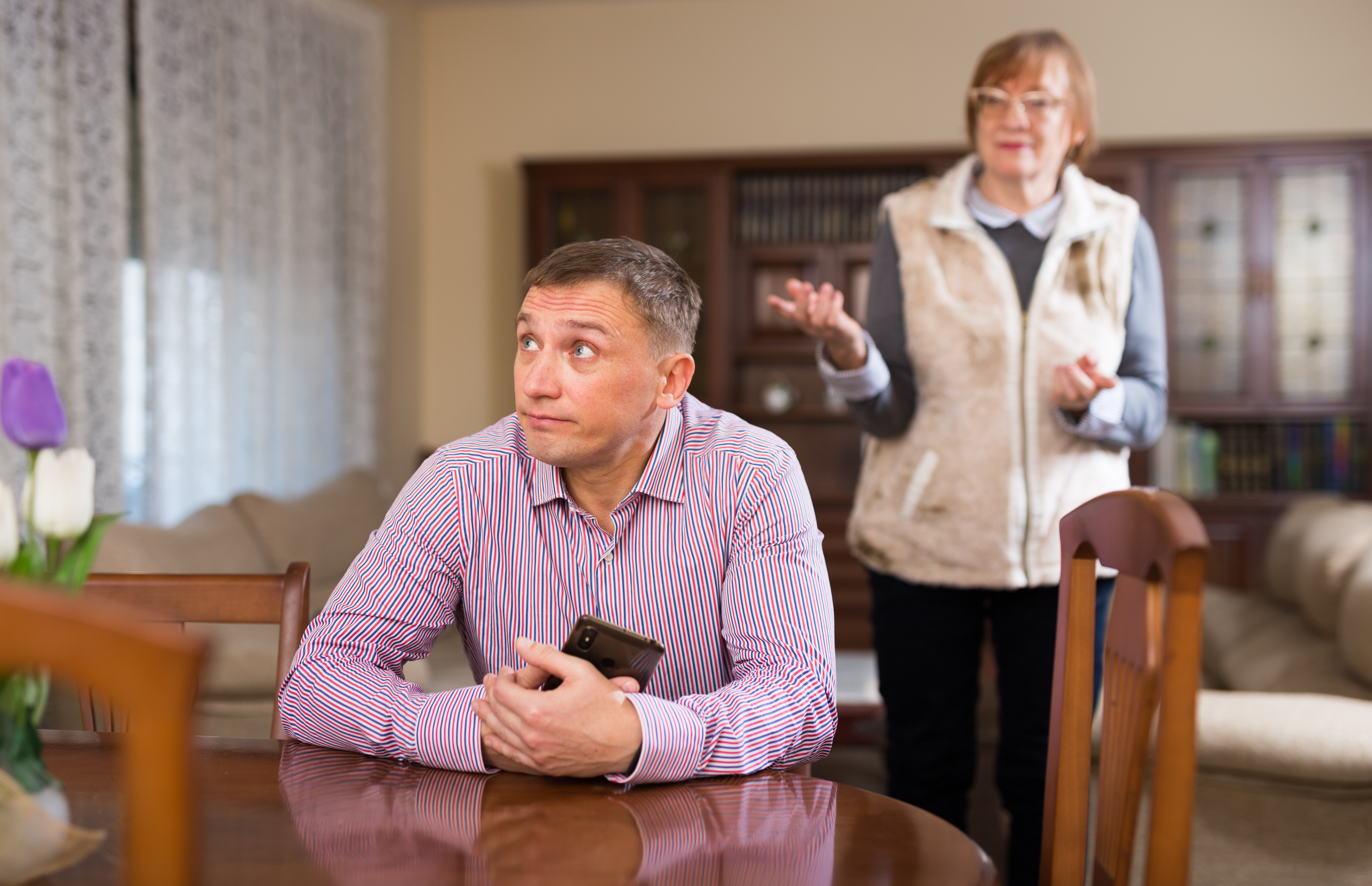 A younger man clutching his phone and looking away while sitting at a table while an older lady tries to speak to him in the background | Source: Shutterstock